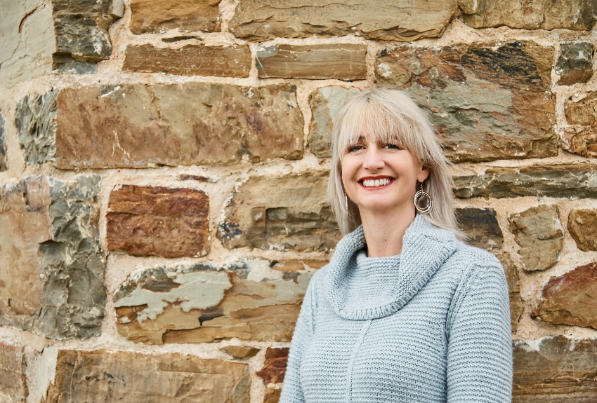 Person wearing a blue jumper and leaning against a brick wall smiling at the camera
